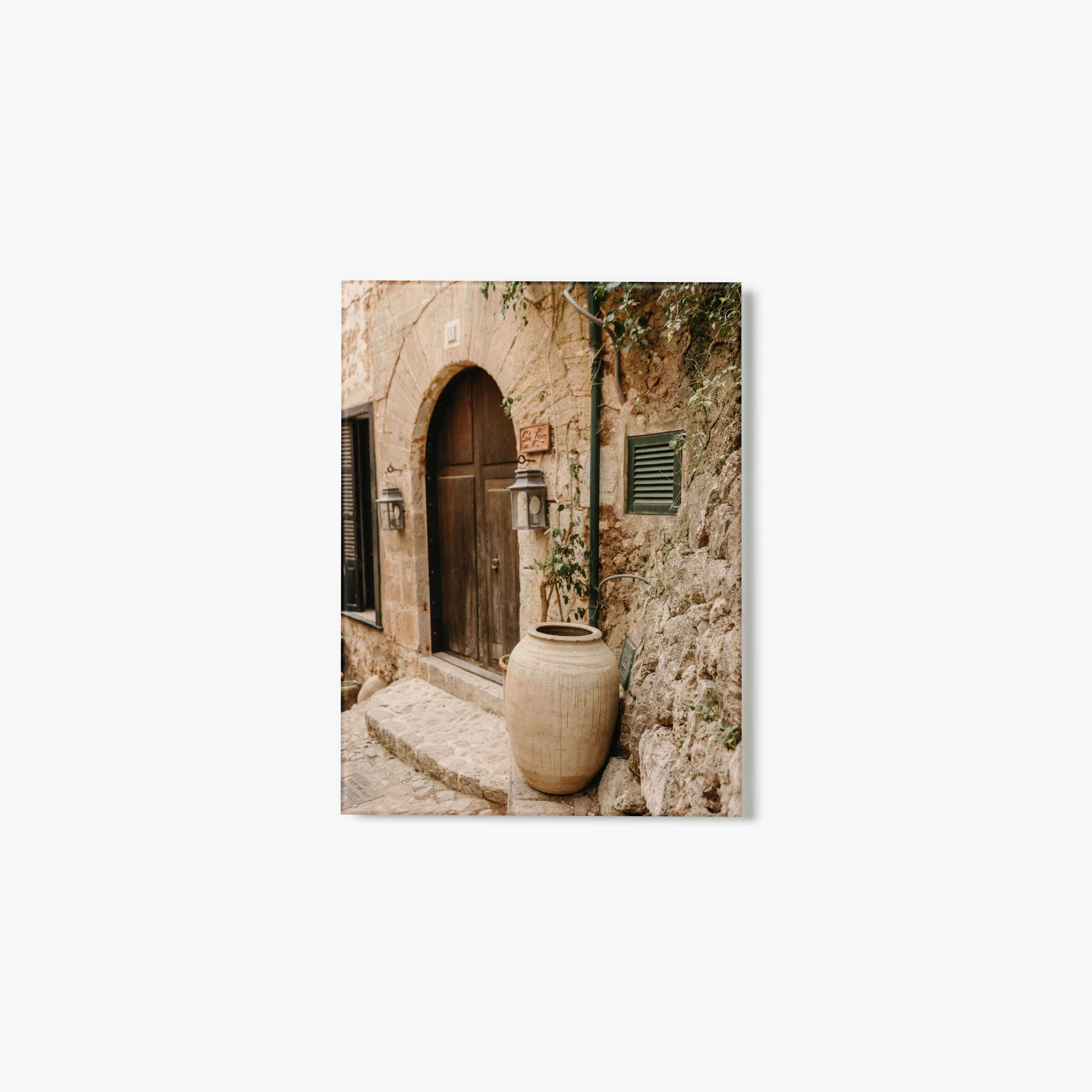 Glass Print of a stone archway with wooden door and large ceramic pot in a narrow alley.