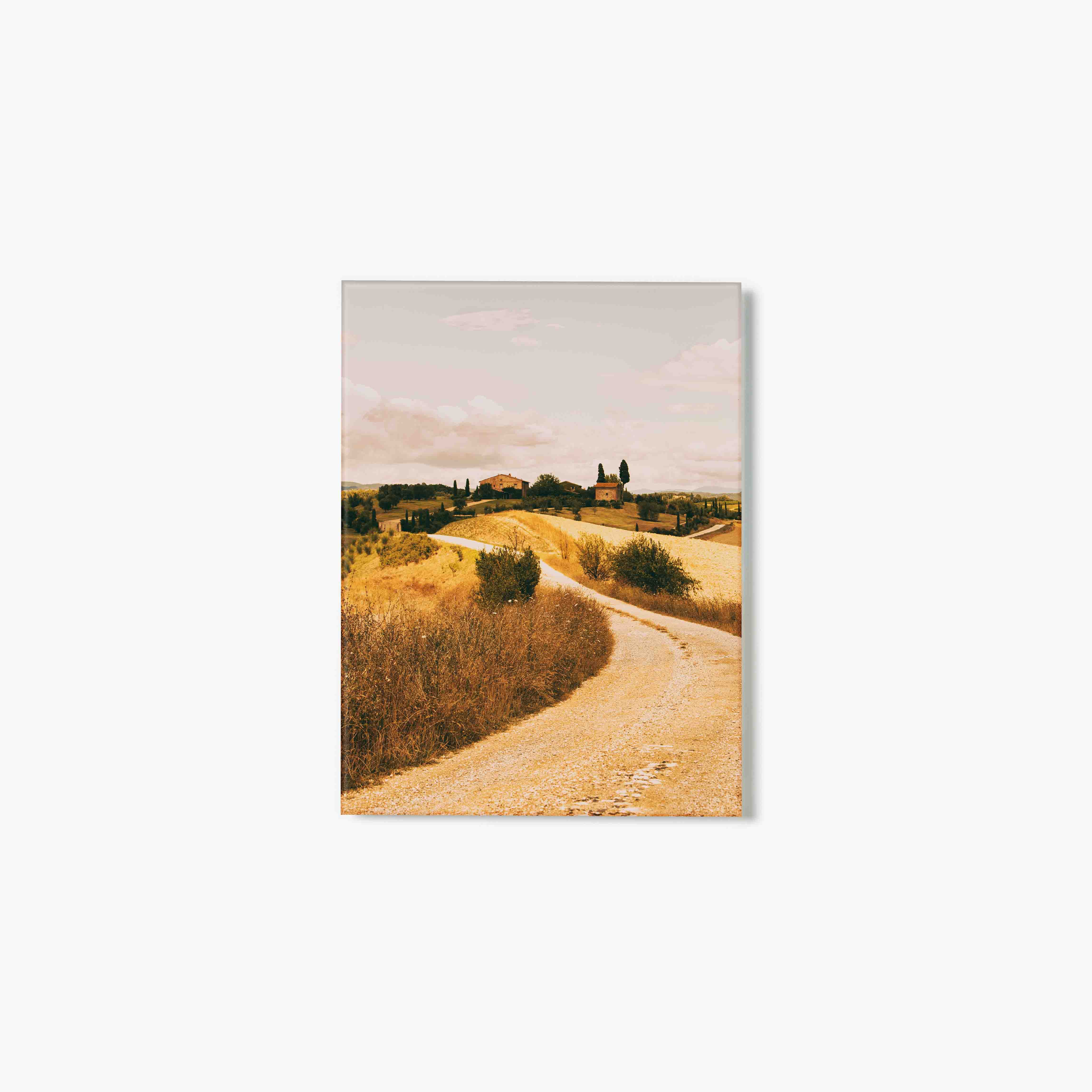 Glass print of winding road through a scenic landscape with rolling hills and buildings.